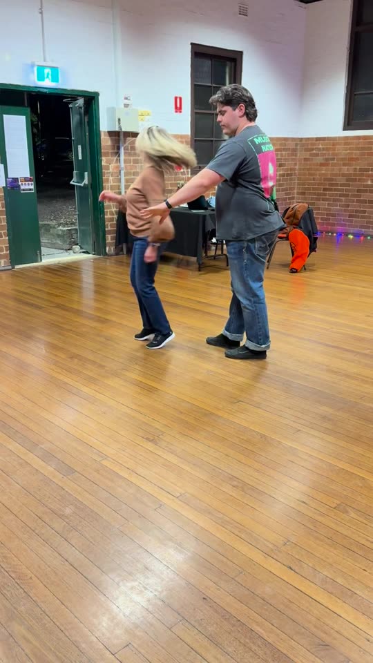 A woman and a man practice a slow partner turn on a wooden floor in a community hall, filmed on a phone.